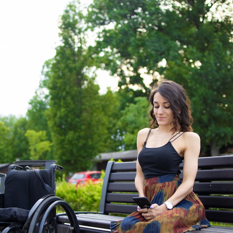 A wheelchair user sits on a bench in a park, looking at her phone.